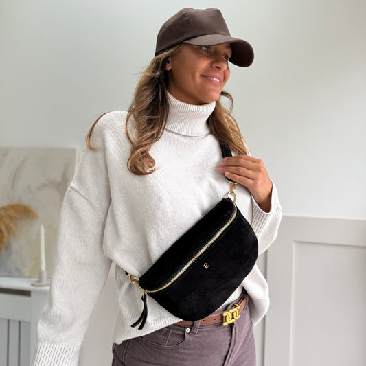 Woman wearing a white sweater, brown cap, and holding a black bag against a white wall.
