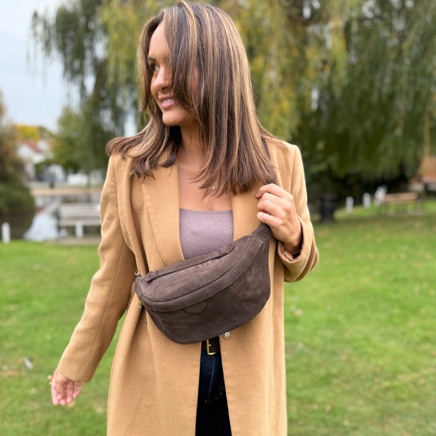 Woman in a beige coat holding a brown bumbag outdoors with greenery in the background