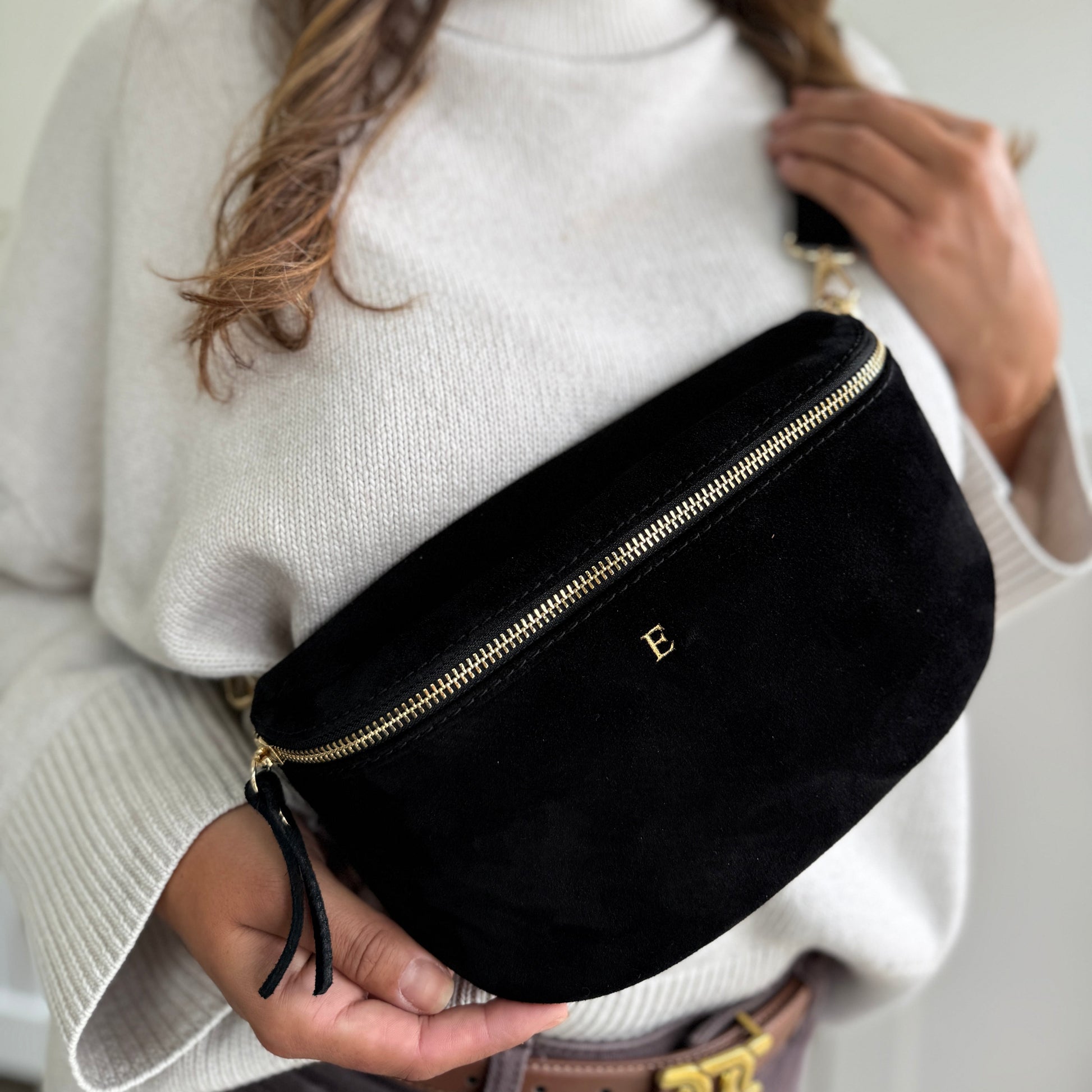 Person wearing a black velvet fanny pack with a gold zipper, holding it in front of a white background.