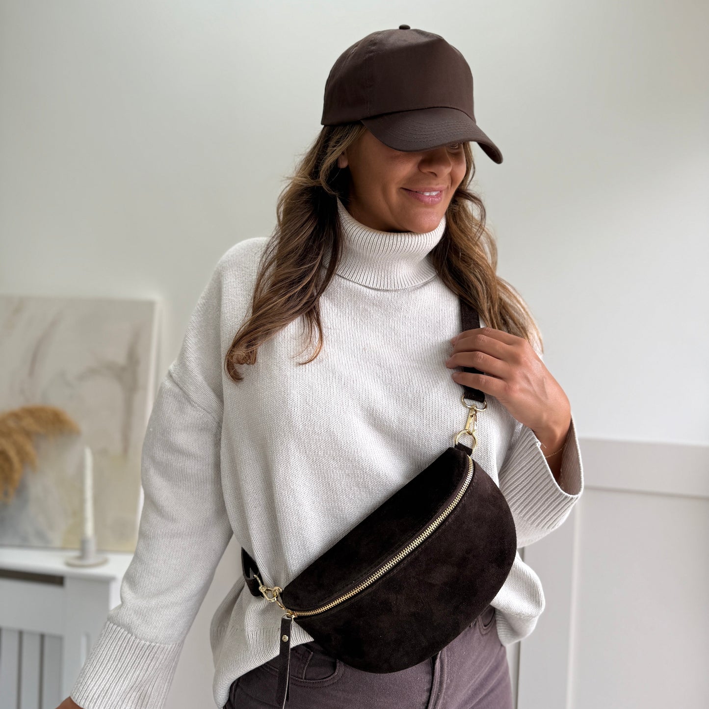 Woman wearing a white turtleneck, brown cap, and brown waist bag indoors.
