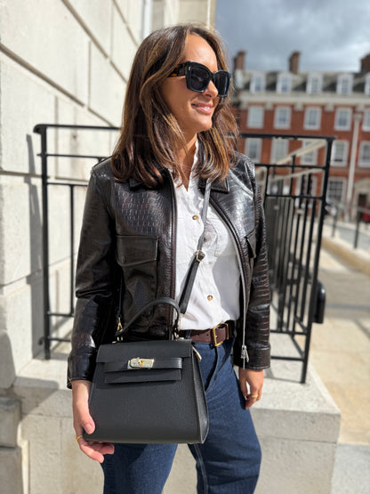 Woman in a black leather jacket and sunglasses holding a black handbag on a city street.