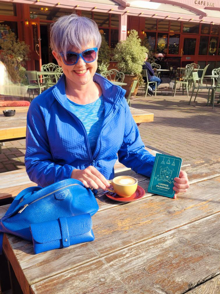 Woman in blue jacket and sunglasses sitting at an outdoor cafe with blue leather bag leather glasses case
