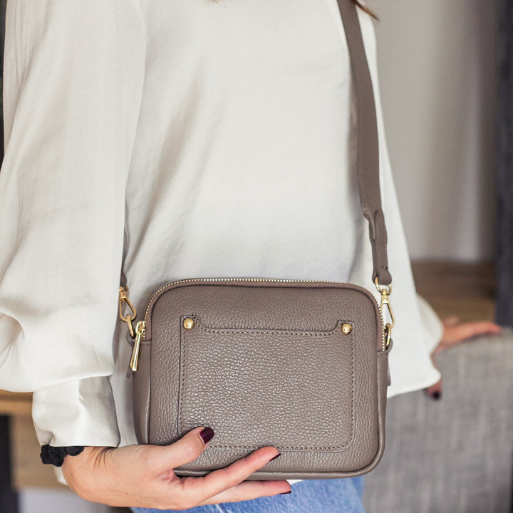 A woman carrying a dark taupe leather double zip crossbody bag with a detachable strap over her shoulder.