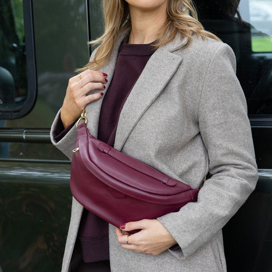 Woman in gray coat holding a burgundywaist bag, standing next to a vehicle.