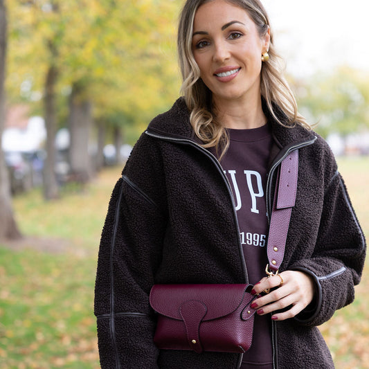 Woman holding a burgundy handbag in an outdoor setting with trees and grass.